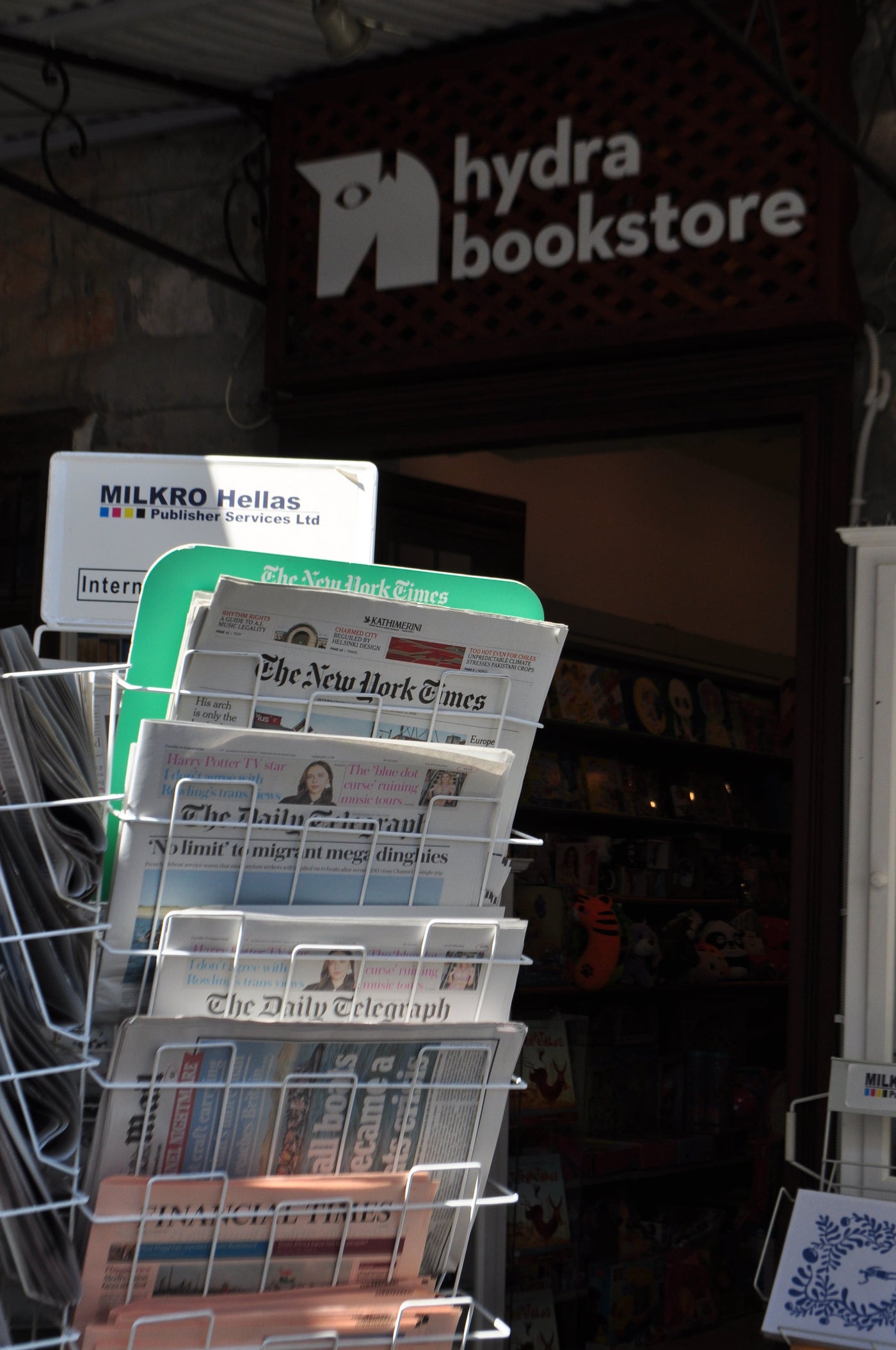 A wire magazine rack filled with newspapers, including The New York Times and The Daily Telegraph, set in front of Hydra Bookstore. Shot by Eleanna Tabouri , creative director of  Nevro Blazer location Hydra 2026