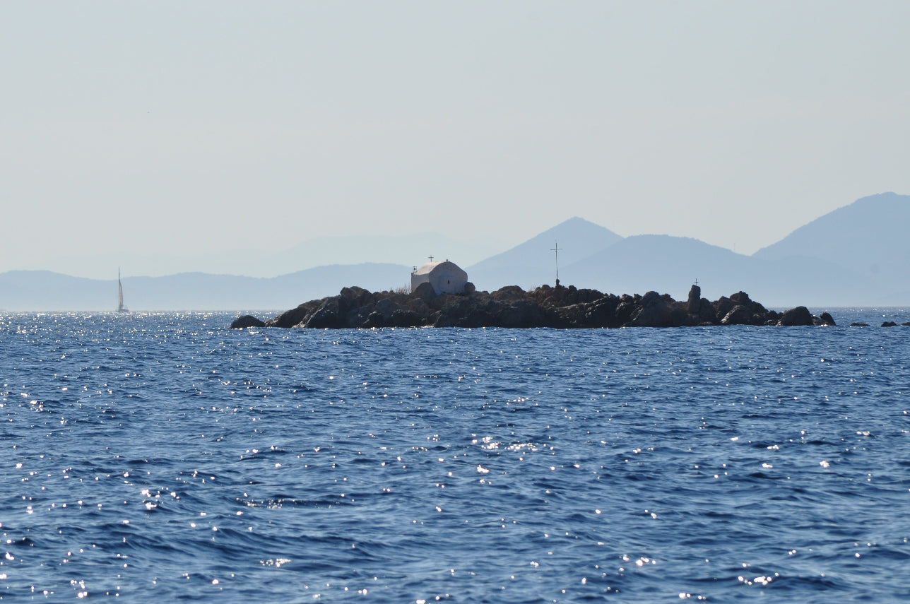 A small rocky island in the blue sea with a white building on top, distant mountains, and a pale hazy sky.
