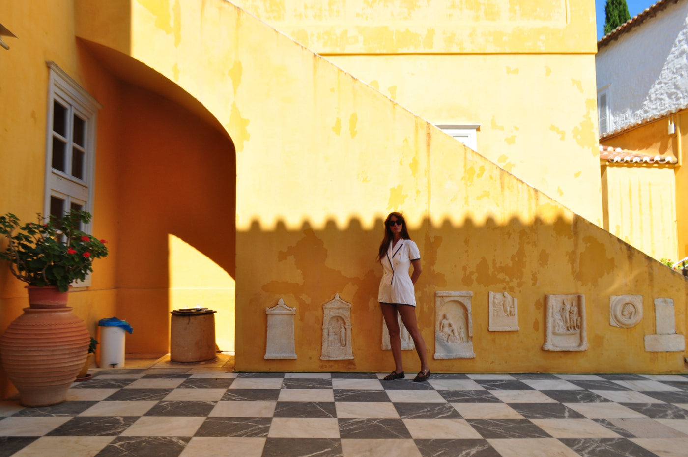 Eleanna Tabouri, Creative Director of Nevro Blazer,  in a vanilla Terry playsuit stands on a black-and-white checkered floor in front of a large yellow building wall with arches, potted plants, and small stone relief sculptures . Location Lazaros Koundouriotis Mansion Museum Hydra Island, Greece