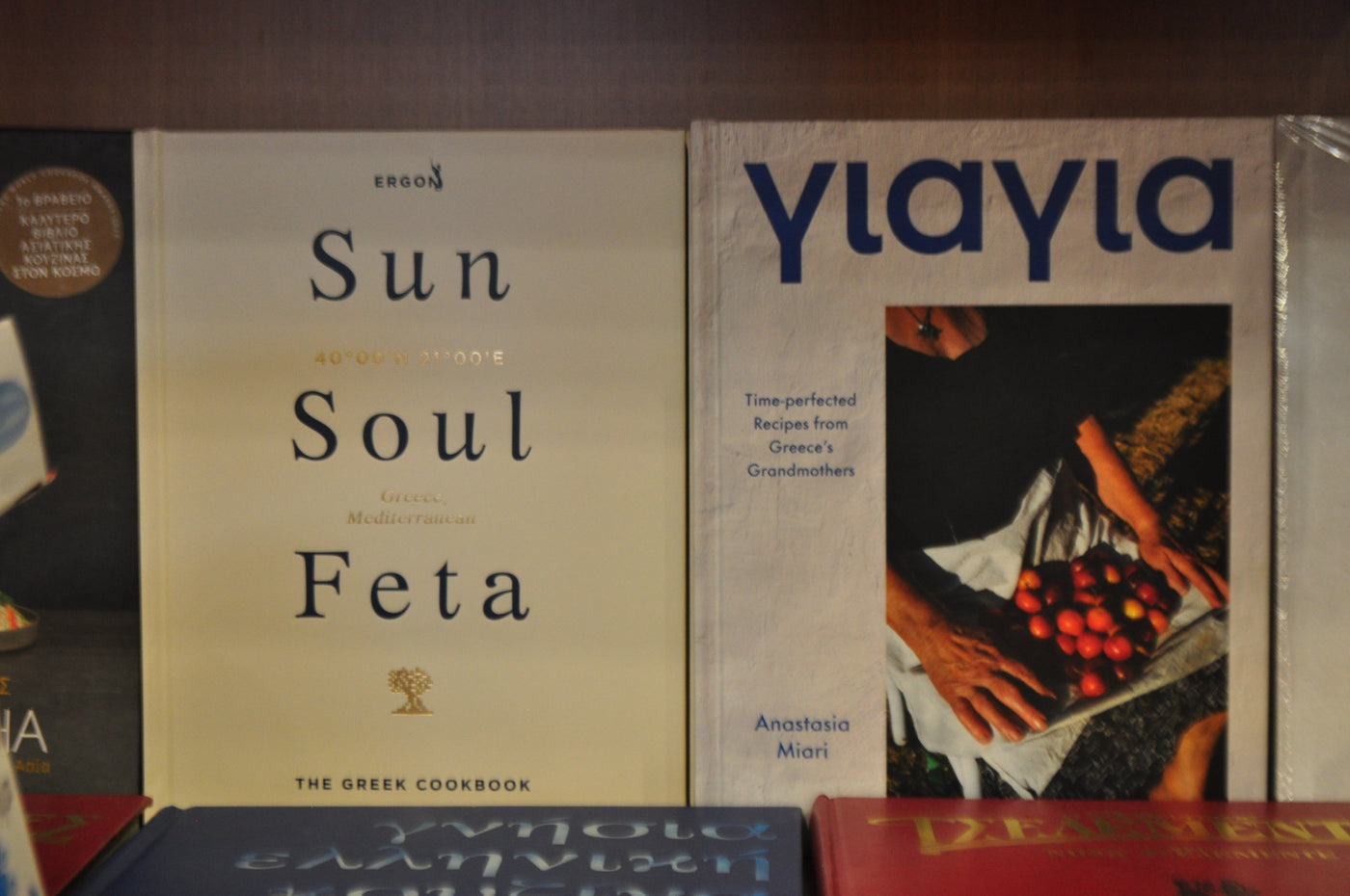 Two cookbooks displayed on a shelf: 'Sun Soul Feta' by Ergon on the left with a cream cover, and 'Ylayia' by Anastasia Mioti on the right with a dark cover featuring a photo of hands holding red olives. Postcards for Hydra 2026 shot by Eleanna Tabouri creative director of Nevro Blazer