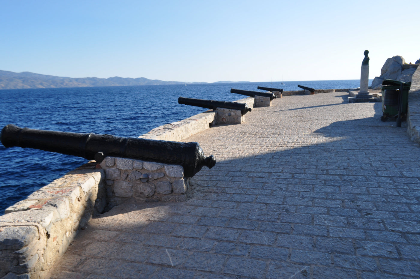 A stone walkway along the edge of a blue sea, Bastions with cannons, location Hydra, Greek Island. A small statue stands near the right side, with distant mountains visible under a clear blue sky. The historic cannons and bastions on the Greek island of Hydra sit along the harbor entrance. Built in the 18th century, these coastal fortifications and rusty chains protected the local fleet and port from the Ottoman-Turkish fleet. Shot by Eleanna Tabouri Creative Director Nevro Blazer