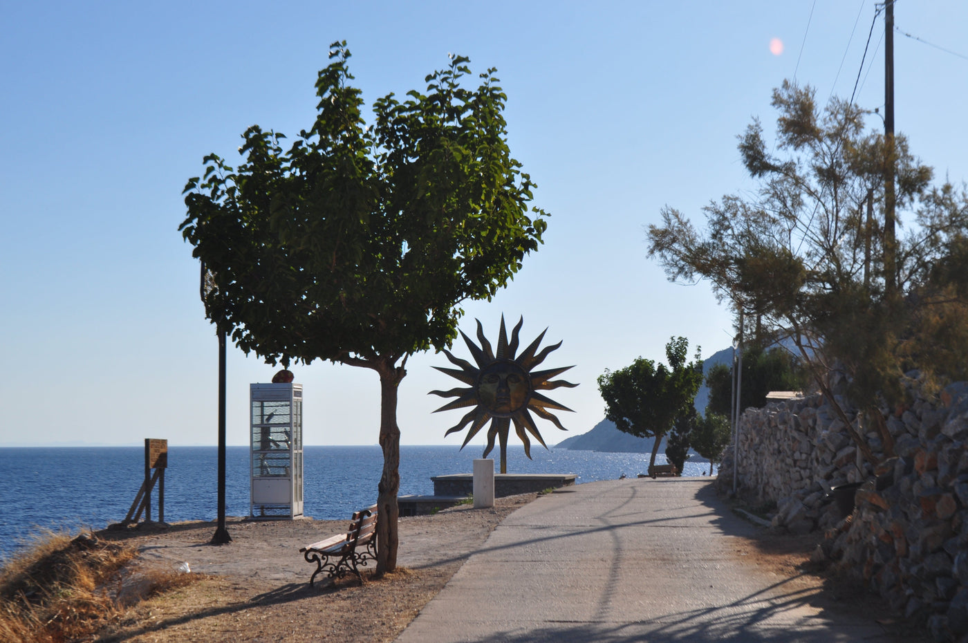 A coastal pathway beside the sea with a large sunburst sculpture, a tree, benches, a stone wall, and distant hills under a clear blue sky. The DESTE Foundation & Jeff Koons: Apollo, a solo exhibition on view at DESTE’s Project Space at the old Slaughterhouse in Hydra.  An installation presenting new sculptures by Jeff Koons along with readymade objects selected by the artist to engage the viewer in a metaphysical dialogue. Shot by Eleanna Tabouri, Creative Director of Nevro Blazer, location Hydra, Greek Isl