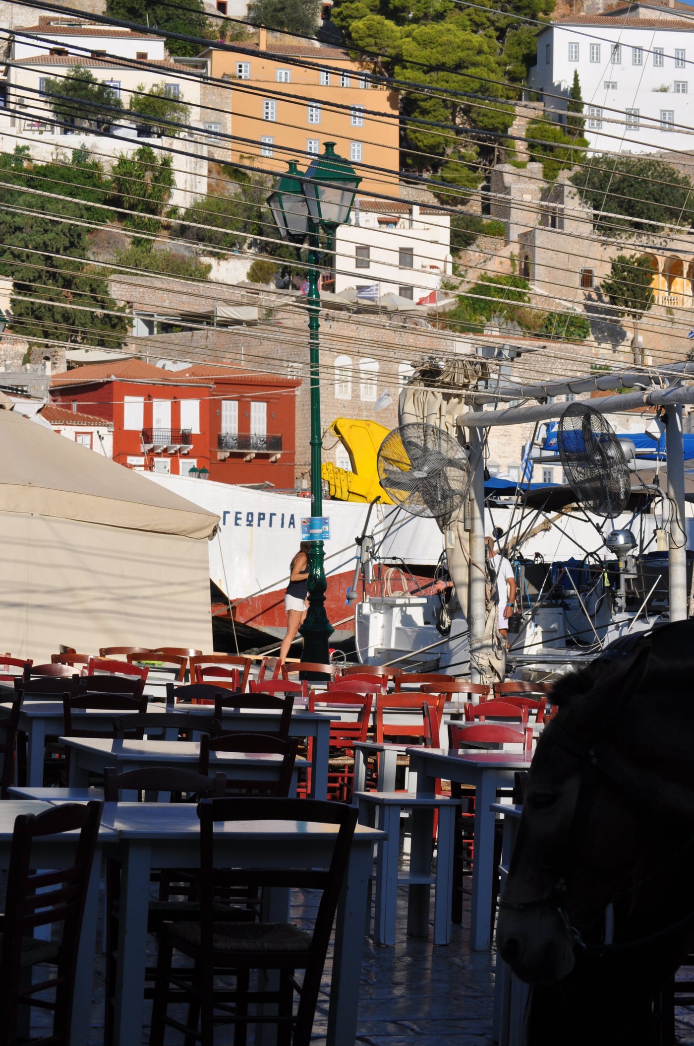 Outdoor dining tables and chairs arranged on a waterfront terrace, with a green lamppost, boats, buildings, and a hillside background. A person is partially visible in the foreground, and the scene has a busy, real-environment feel. Shot by Eleanna Tabouri Creative Director of Nevro Blazer in Hydra 2026