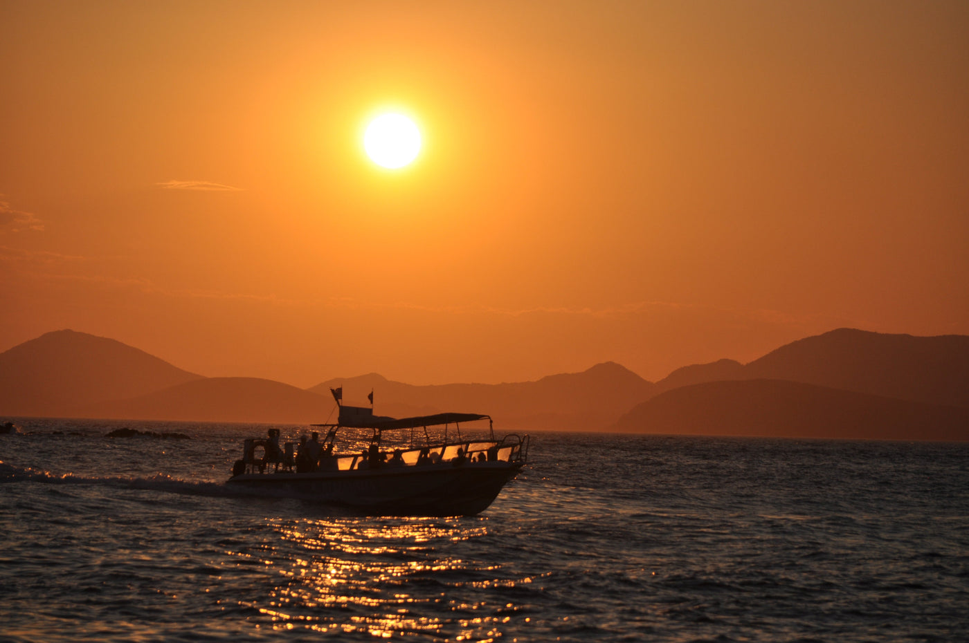 A sunset seascape with a small boat silhouetted against a glowing orange sky and distant mountains. Shot by Eleanna Tabouri, Creative Director of Nevro Blazer. Location Hydra 2026.