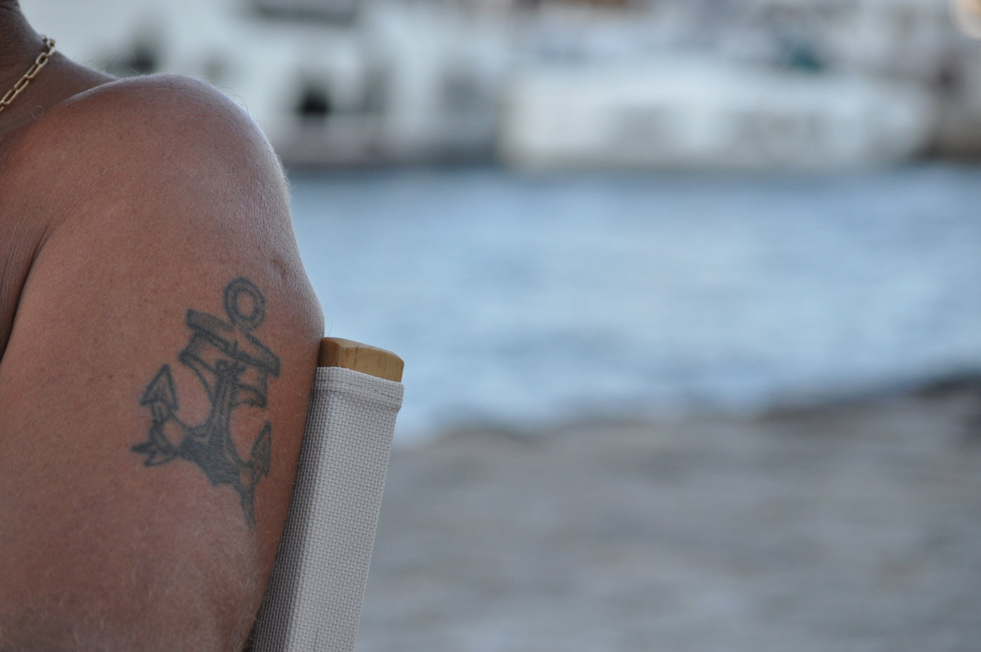 A close-up of a person's bare shoulder featuring a black anchor tattoo, with a blurred beach and water background. Shot byEleanna Tabouri, Creative Director of Nevro Blazer, location Hydra 2026.