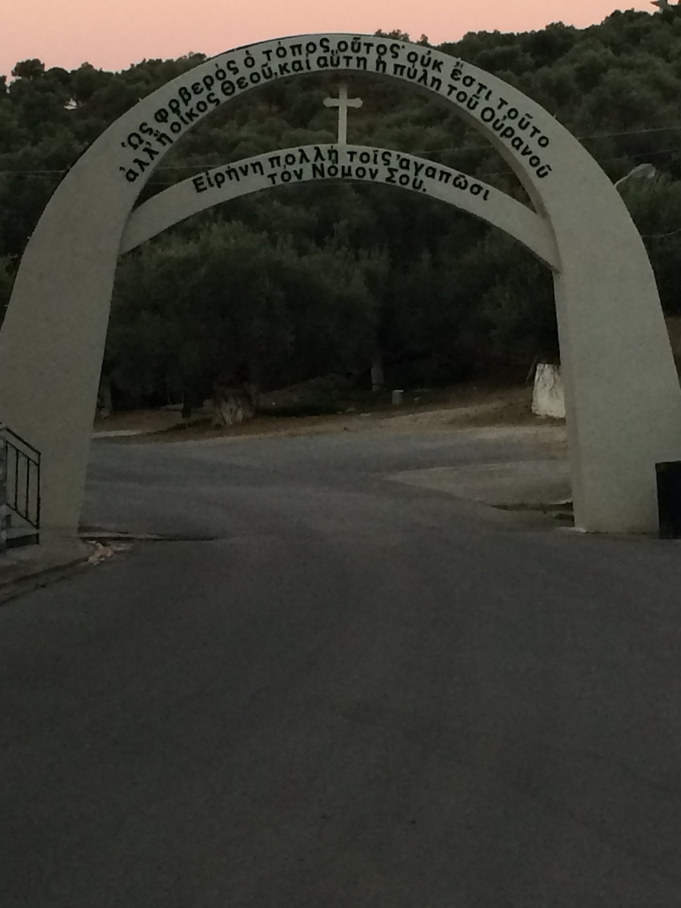 A large concrete archway with Greek text and a small cross at the top, opening onto a road with dark trees in the background at dusk. monastery_saintrafael_lesvos