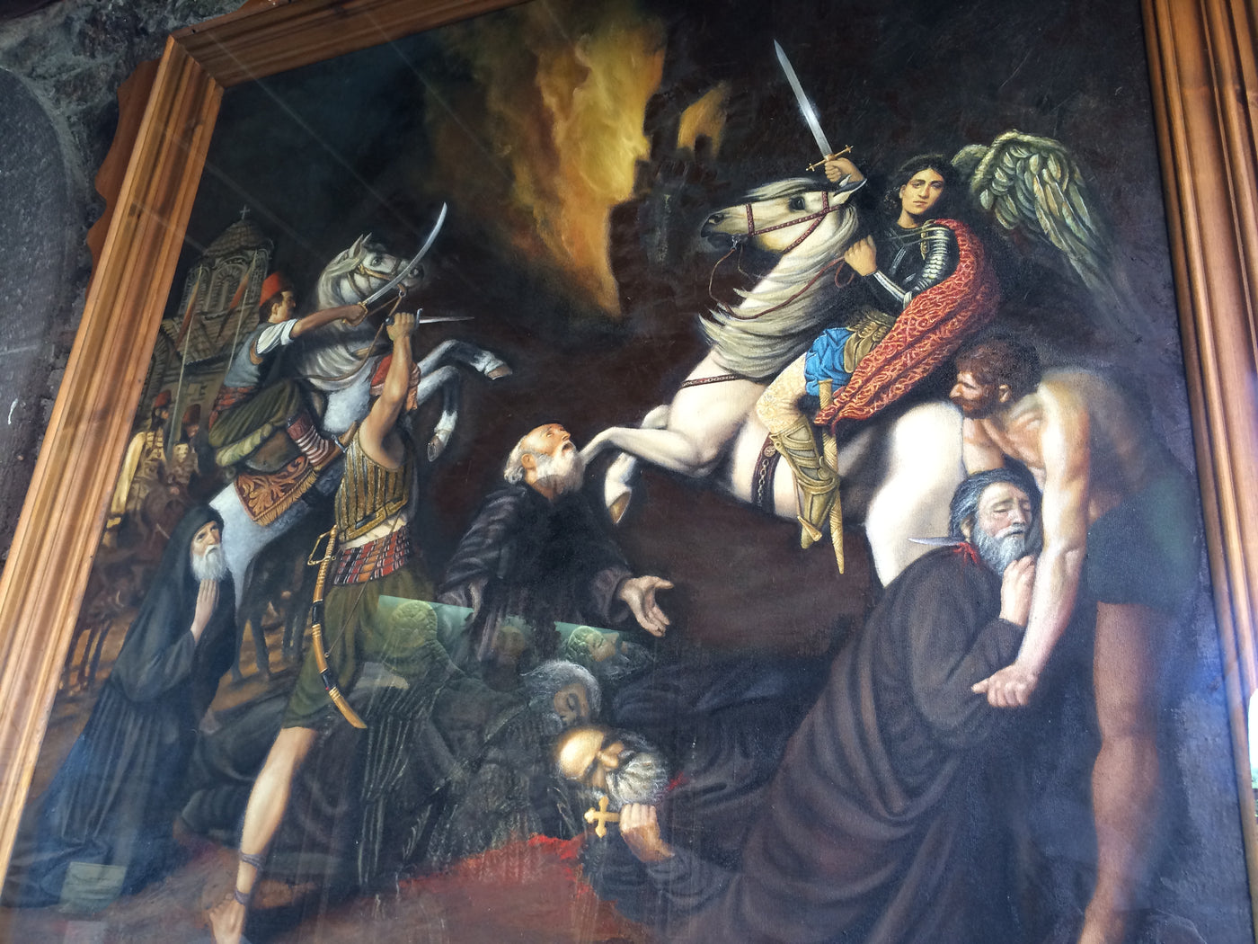 A large framed historical painting depicting a dramatic battle scene with a mounted warrior holding a sword, figures in dark robes, flames in the background, and a wooden frame bordering the artwork. archaengel Michael monastery lesvos