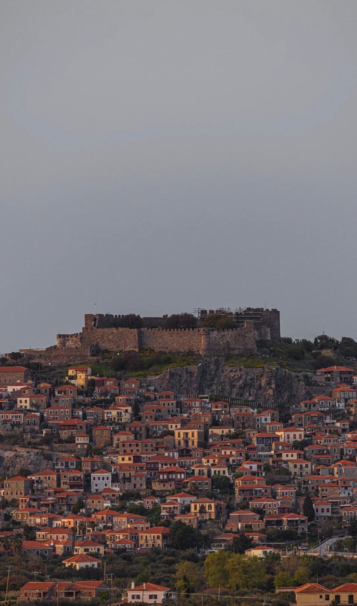 A scenic view of a hillside town with dense orange-roofed houses and a large stone fortress perched atop a rocky cliff under a cloudy gray sky. lesvos