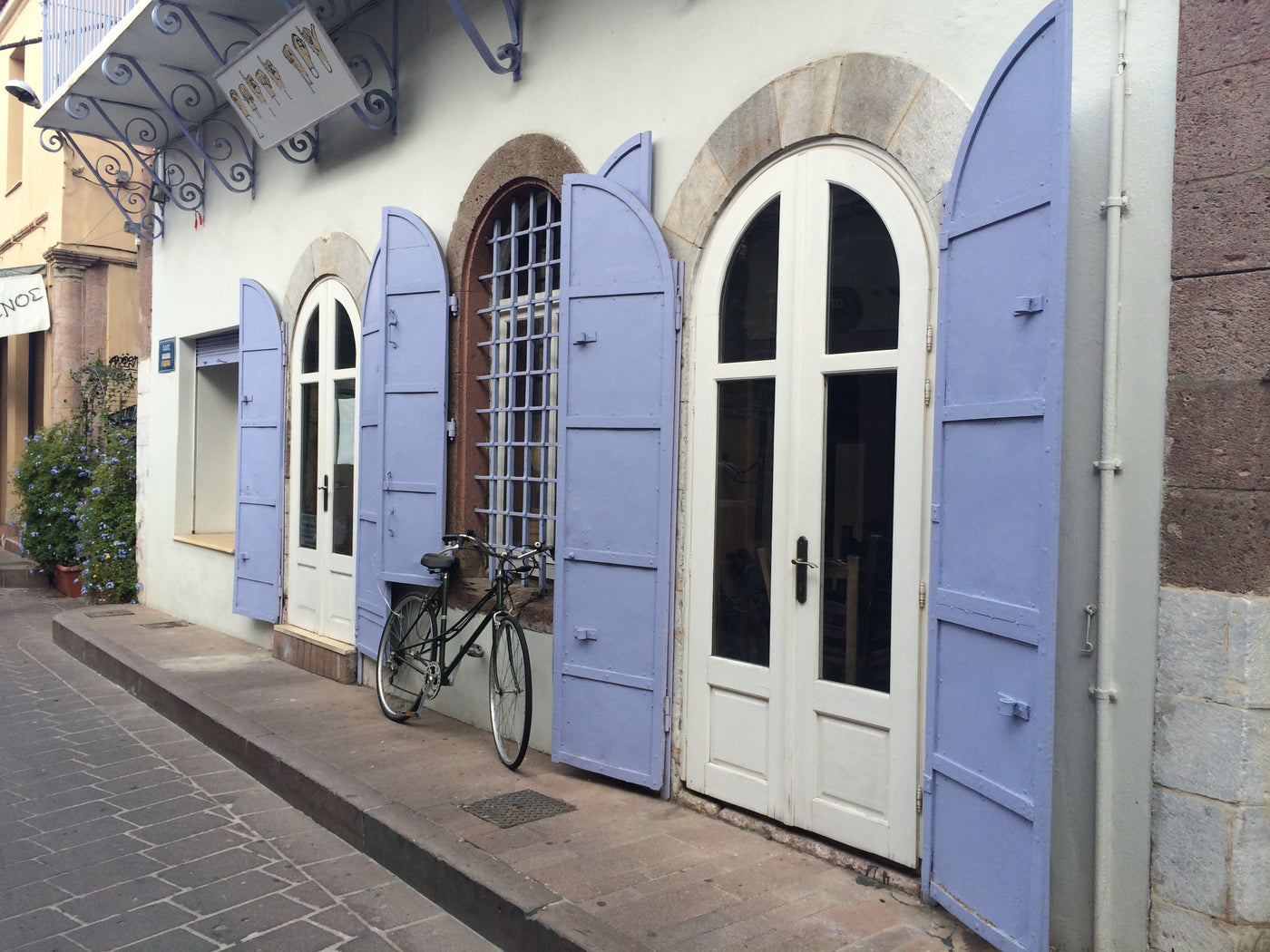 A street view of a building facade with white arched double doors and windows framed by lavender-blue shutters, alongside a sidewalk, cobblestone street, and a parked bicycle. lesvos