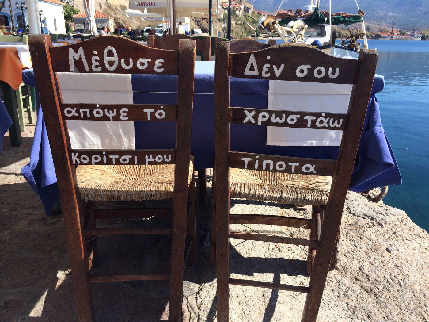 Two wooden dining chairs at an outdoor seaside table, decorated with Greek flag panels and white Greek text, set against a rocky shoreline with blue water and boats in the background. lesvos