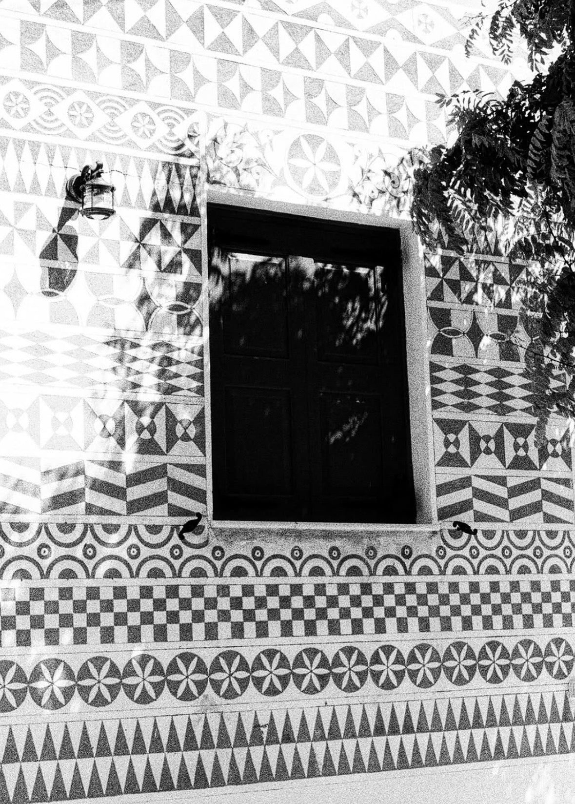 Black and white photograph of a building facade featuring intricate geometric patterned stucco tiles surrounding a dark window, with leafy tree branches casting shadows across the wall. Pyrgi Chios
