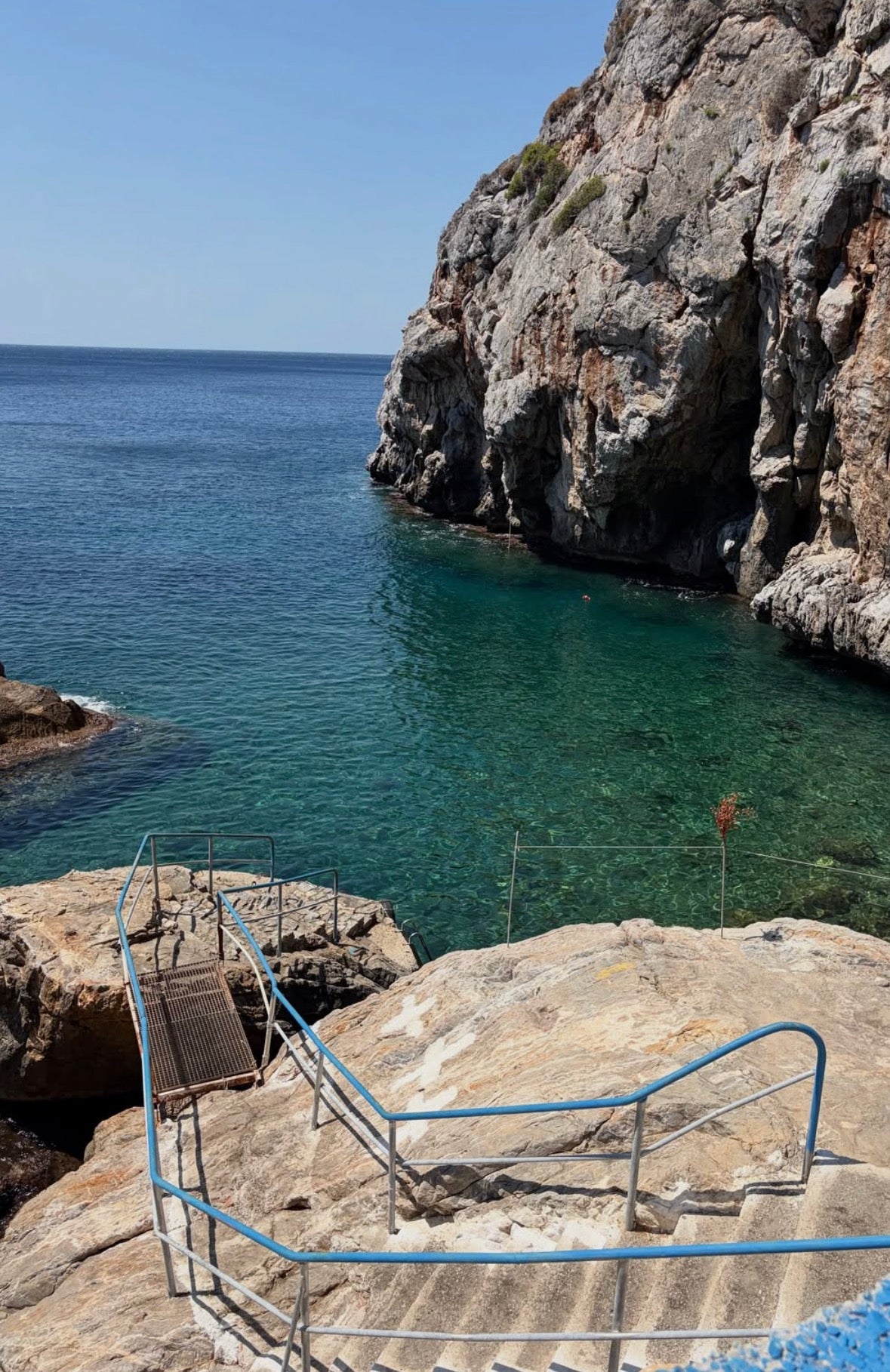 A scenic coastal view with clear turquoise water, steep rocky cliffs, and a stone staircase with blue railings leading down to the sea under a blue sky. PANAGIA KRIFTI_LESVOS